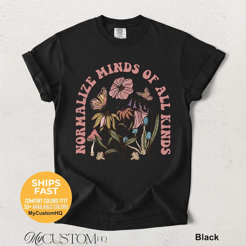 May include: A black short-sleeve t-shirt featuring a graphic print of flowers, butterflies, and mushrooms arranged in an arch. The text NORMALIZE MINDS OF ALL KINDS is printed in a light pink arched font around the central botanical illustration.