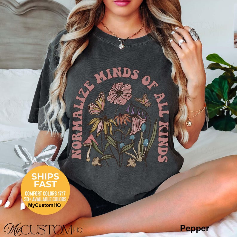 May include: A charcoal gray short-sleeve t-shirt featuring a graphic print of various flowers, mushrooms, and a butterfly. The text "NORMALIZE MINDS OF ALL KINDS" is printed in a curved, light pink font around the central floral illustration.