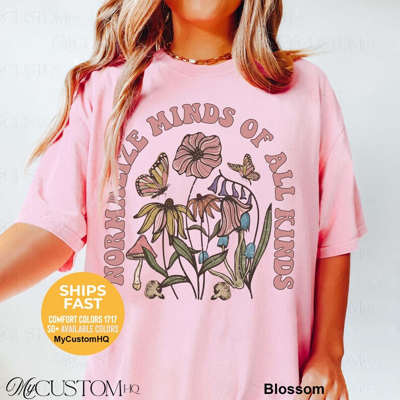 May include: A light pink t-shirt featuring a graphic print of various flowers, mushrooms, and butterflies. The text "NORMALIZE MINDS OF ALL KINDS" is arched over the floral design in a muted brown font. The shirt has short sleeves and a crew neckline.