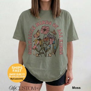May include: A moss green short-sleeve t-shirt featuring a graphic print of various flowers, mushrooms, and butterflies. The text "NORMALIZE MINDS OF ALL KINDS" is printed in a curved arch around the central botanical illustration in pink letters.