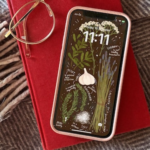 May include: A red book with gold-rimmed glasses and a smartphone with a botanical illustration. The phone displays the time 11:11. The items rest on a gray herringbone patterned blanket, with a cream-colored fringe on the left.
