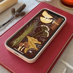 May include: A smartphone with a decorative screen displaying illustrations of herbs, mushrooms, and lemon slices, resting on a red book. The phone's screen also shows the time 11:11. Additional items include candles, a cup, and a lit candle.