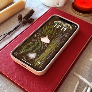 May include: A smartphone with a light pink case displaying a botanical illustration of herbs, garlic, and salt, resting on a red book. The screen shows the time 11:11. Other items include candles, a cup, and a dried flower.