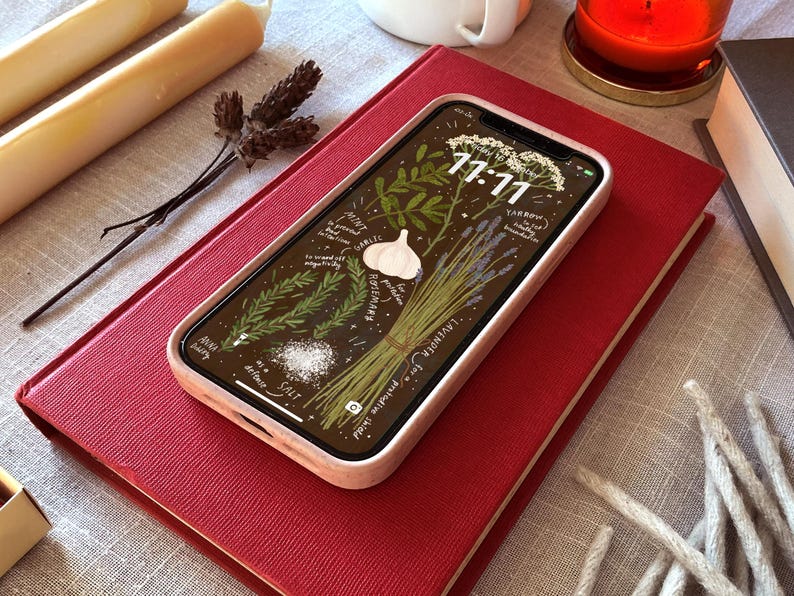 May include: A smartphone with a light pink case displaying a botanical illustration of herbs, garlic, and salt, resting on a red book. The screen shows the time 11:11. Other items include candles, a cup, and a dried flower.