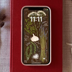 May include: A smartphone displays a botanical illustration wallpaper with herbs and text. The screen shows a brown background with drawings of mint, garlic, rosemary, lavender, and other plants. The time 11:11 is displayed. The phone is on a red book.