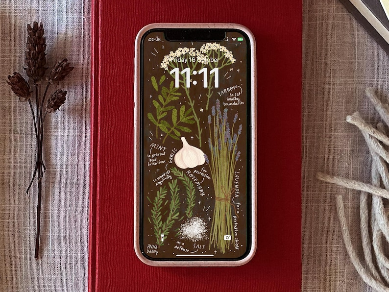 May include: A smartphone displays a botanical illustration wallpaper with herbs and text. The screen shows a brown background with drawings of mint, garlic, rosemary, lavender, and other plants. The time 11:11 is displayed. The phone is on a red book.