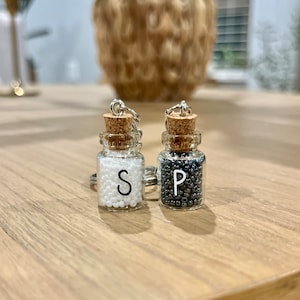 May include: Two small glass jars with cork tops and silver keychains. The jars are filled with white and black beads and labeled "S" and "P".