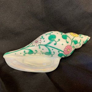 May include: A white ceramic seashell with a green, white, pink, and yellow floral design. The shell is a decorative object.