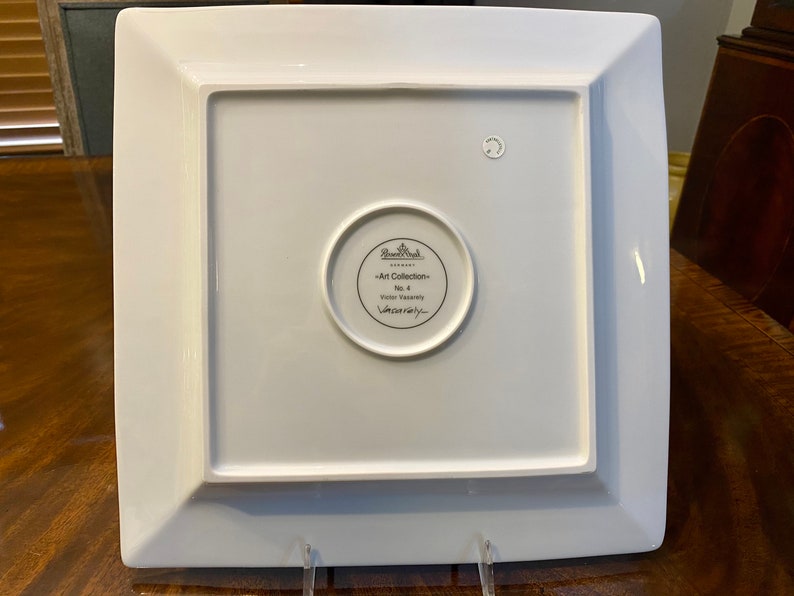 Puede incluir: Plato cuadrado de cer&aacute;mica blanca con un borde elevado y un logotipo circular en el centro. El logotipo dice "Rosenthal -Art Collection- No. 4 Versace"