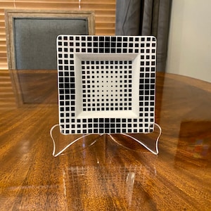 Puede incluir: Un plato cuadrado de cer&aacute;mica blanco y negro con un patr&oacute;n geom&eacute;trico. El plato est&aacute; sobre un soporte de acr&iacute;lico transparente.