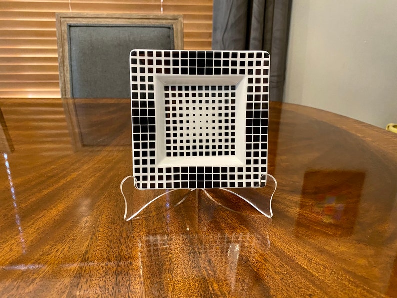 Puede incluir: Un plato cuadrado de cer&aacute;mica blanco y negro con un patr&oacute;n geom&eacute;trico. El plato est&aacute; sobre un soporte de acr&iacute;lico transparente.