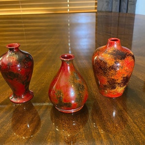 Puede incluir: Tres pequeños jarrones de cerámica roja y negra con un esmalte moteado. Los jarrones están dispuestos en fila sobre una superficie de madera.