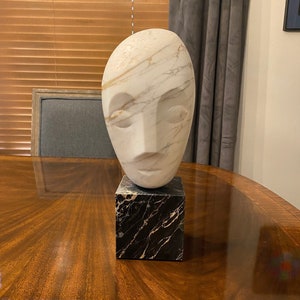 May include: White marble sculpture of a face on a black marble base.