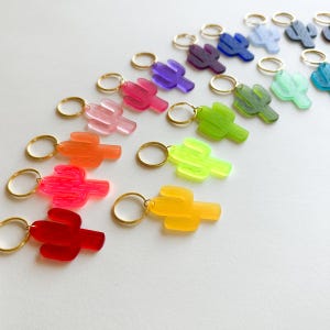 May include: A collection of translucent cactus-shaped charms in a variety of vibrant colors, each attached to a small gold-toned metal ring. The assortment includes red, orange, yellow, pink, green, blue, and purple acrylic cactus pendants.