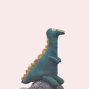 May include: A green knitted dinosaur toy with a yellow spiky back, sitting on a rock.