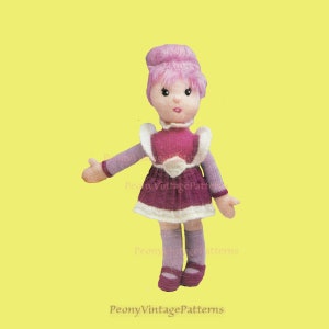 Puede incluir: Una muñeca de punto con pelo rosa, un vestido morado y ribetes blancos. La muñeca lleva calcetines y zapatos morados.