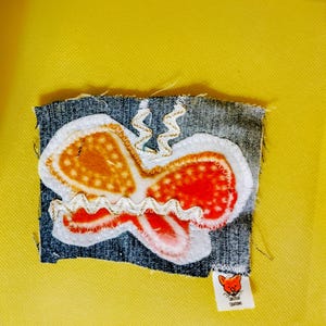 Puede incluir: Un parche rectangular de tela vaquera con una aplicación de mariposa en capas en tonos naranja, rojo y blanco. La mariposa está acentuada con ribete de zigzag blanco y dorado. Una pequeña etiqueta blanca muestra un icono de zorro y el texto "CHEZYCAT CREATIONS".