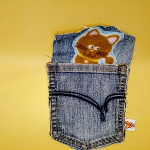 May include: A blue denim fabric pocket featuring a brown cat patch with pink ears and a yellow collar. The pocket has a black stitched decorative line across the front and a small white rectangular tag with a cat illustration on the side edge.