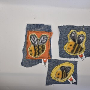 May include: Three fabric patches featuring bee illustrations on blue denim scraps. Each patch has a yellow or orange border with zigzag stitching. The bees are yellow and black with grey wings. Small white tags with a red fox logo are attached.