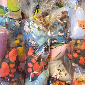 Puede incluir: Una colecci&oacute;n de juguetes de tela con tem&aacute;tica de gatos, cada uno empaquetado individualmente en pl&aacute;stico transparente. Los juguetes presentan varios patrones, incluyendo ilustraciones de gatos, huellas de patas y espinas de pescado. Los colores son vibrantes, con azules, naranjas y rojos.