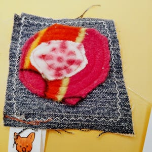 May include: A square dark blue denim fabric patch featuring a circular layered applique in shades of red, orange, and white. The patch is finished with a white zigzag stitch border and includes a small white tag with a cat illustration and the text CHEEZECAT CREATIONS.