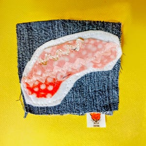 Könnte beinhalten: Ein Jeans-Patch mit einem Filz-Applikation mit einem rosa-weißen Design mit roten Tupfen und silbernen Fadenakzenten. Der Patch befindet sich auf einem gelben Hintergrund, mit einem kleinen Etikett mit der Aufschrift "Sweetcat Creations".