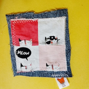 May include: A square fabric patch featuring a four-panel grid with illustrations of cats. The panels are red, white, and light pink, bordered by dark blue denim with frayed edges. One panel includes a black speech bubble with the text MEOW.