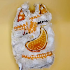 May include: A plush, white and gray oven mitt with orange accents. The mitt features orange and white patterned patches, a large orange paisley design, and black stitching. A small white label with text is visible at the bottom.