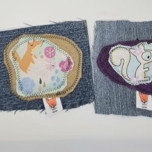 May include: Two rectangular denim fabric patches featuring sewn-on animal designs. The left patch displays a fox and floral print on a tan background. The right patch features a squirrel on a light blue background with a dark purple border.