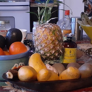 Puede incluir: Naturaleza muerta con pi&ntilde;a, naranjas, aguacates y mangos. Tambi&eacute;n hay una botella de t&oacute;nico y un frasco de Moringa. La fruta est&aacute; dispuesta en cuencos y una bandeja de madera.