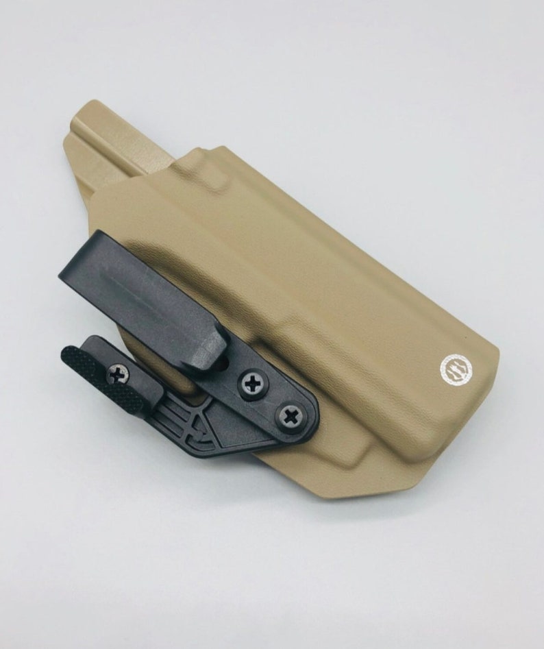 May include: A tan leather holster with a black clip and a black adjustment lever. The holster has a small, round logo on the side.