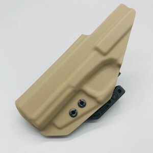 May include: A tan-colored holster with a black attachment. The holster has two black screws on the side.