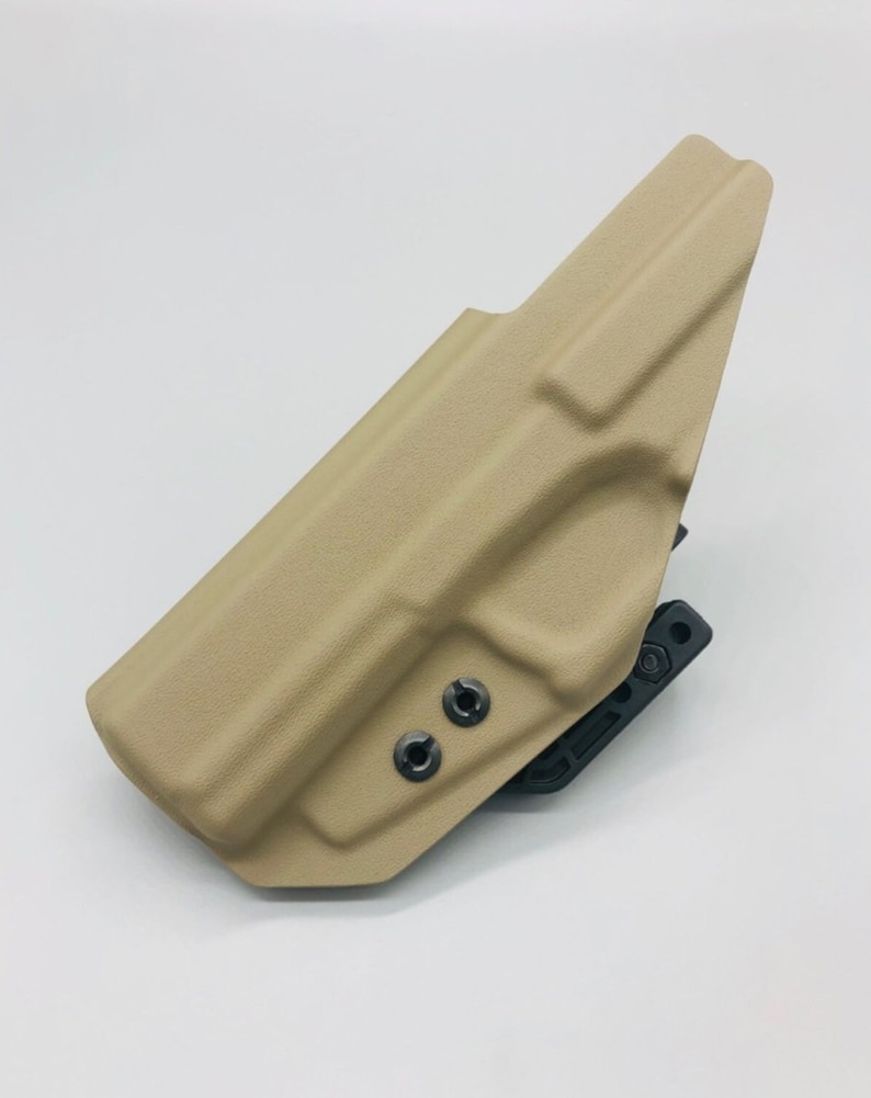 May include: A tan-colored holster with a black attachment. The holster has two black screws on the side.