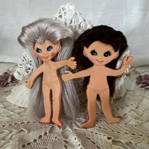 May include: Two vintage doll figures with painted faces, blue eyes, and long hair. One doll has gray hair, the other has dark brown hair. Both dolls are nude and have outstretched arms, standing on a lace surface.