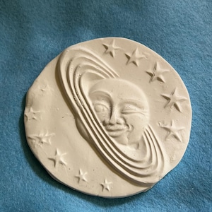 May include: A circular, off-white plaster relief featuring a smiling face surrounded by a ringed design, possibly representing a planet. Stars are evenly spaced around the outer edge. The artwork is set against a blue background.
