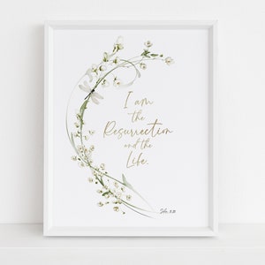 Könnte beinhalten: Weiß gerahmter Kunstdruck mit floralem Design, Libelle und dem Text "I am the Resurrection and the Life". Das Kunstwerk ist im Aquarellstil gehalten, mit goldfarbenen Buchstaben und zarten weißen Blüten.