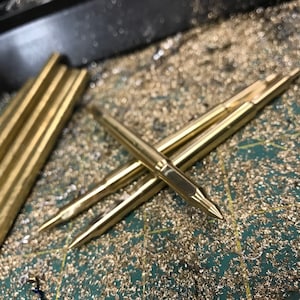 May include: Close-up of several gold-colored metal rods with pointed ends. The rods are lying on a green surface covered in gold-colored metal shavings.