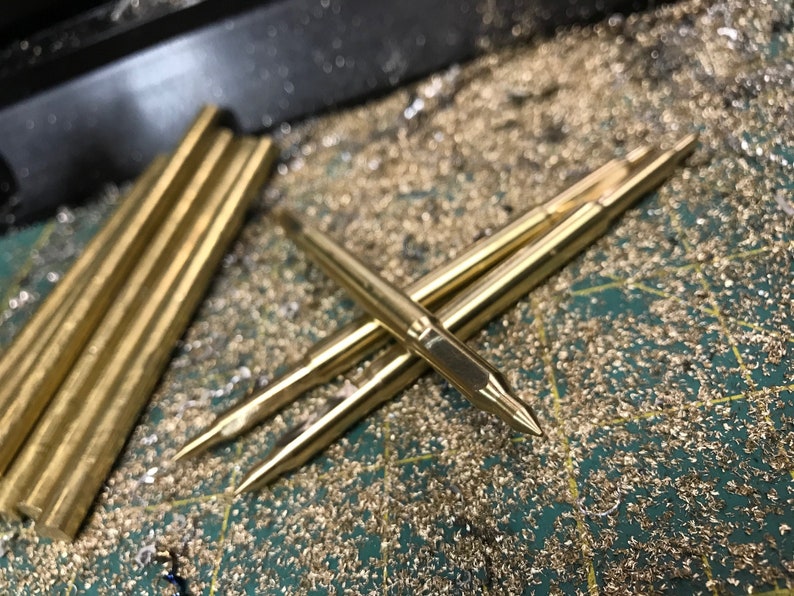 May include: Close-up of several gold-colored metal rods with pointed ends. The rods are lying on a green surface covered in gold-colored metal shavings.
