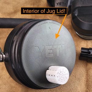 May include: Close-up of the interior of a gray Yeti jug lid. The lid has a black handle and the Yeti logo is visible. The lid is sitting on a brown surface.