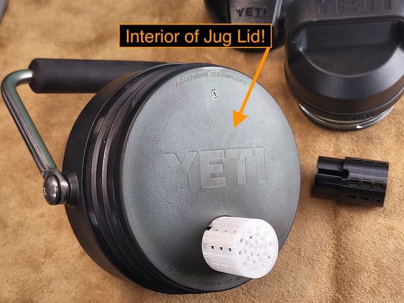 May include: Close-up of the interior of a gray Yeti jug lid. The lid has a black handle and the Yeti logo is visible. The lid is sitting on a brown surface.