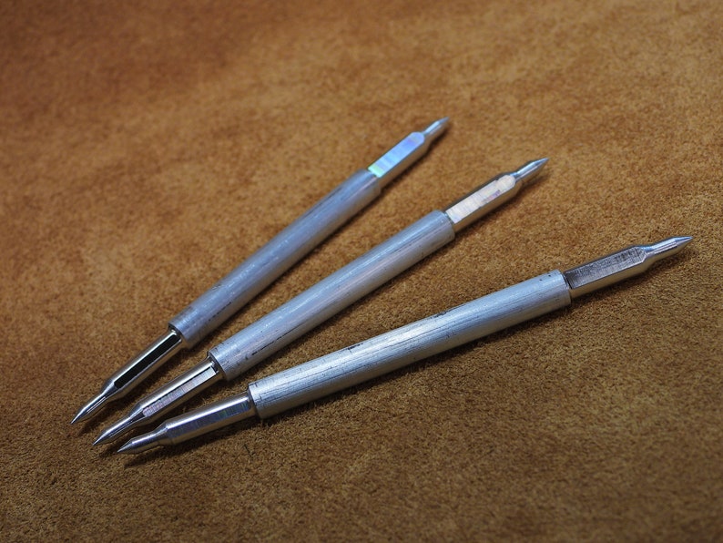 May include: Three silver metal tools with pointed tips, resembling awls or leatherworking tools, arranged on a brown leather surface.