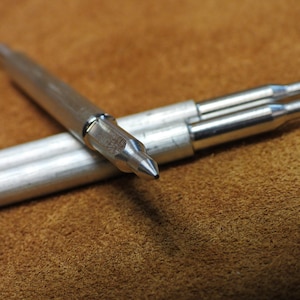 May include: Three silver metal pen-like objects with pointed tips, lying on a brown suede surface.