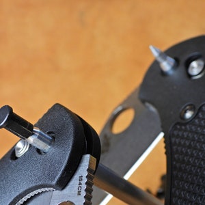 May include: Close-up of a black knife sharpener with a silver metal rod and a black handle. The sharpener has a small silver screw and the text "15.4CM" is printed on the side.