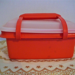 May include: A red plastic storage container with a white lid and two handles. The container is sitting on a white and gold lace tablecloth.
