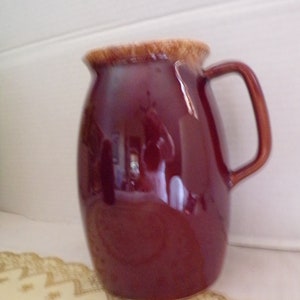 May include: A maroon ceramic pitcher with a brown rim and a handle. The pitcher has a glossy finish and is sitting on a white surface.