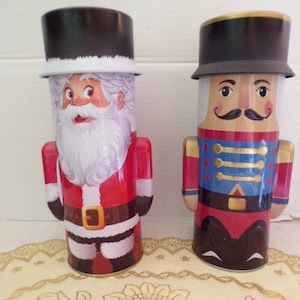 May include: Two vintage Christmas themed tin canisters. One features a Santa Claus design with a red suit, white beard, and black hat. The other features a Nutcracker design with a red and blue uniform, black hat, and a mustache.