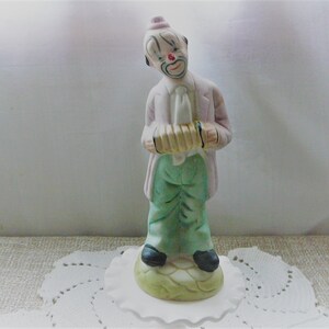May include: A porcelain figurine of a clown playing an accordion. The clown is wearing a pink hat, a white shirt, a green vest, and green pants. The clown has a painted-on red nose and a white face with black eyes and a black mouth.