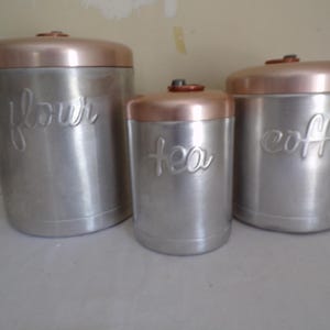 May include: Set of three vintage metal canisters with copper lids. The canisters are labeled "flour", "tea", and "coffee".