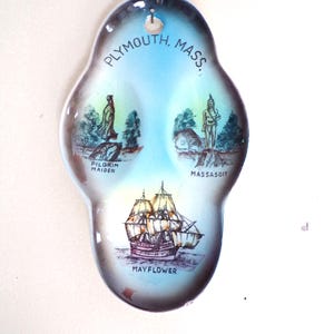 Puede incluir: Un plato de cerámica azul y blanco con imágenes de la Pilgrim Maiden, Massasoit y el Mayflower. El plato tiene forma de lágrima y tiene un agujero en la parte superior para colgarlo. El texto "PLYMOUTH, MASS." está impreso alrededor del borde superior del plato.