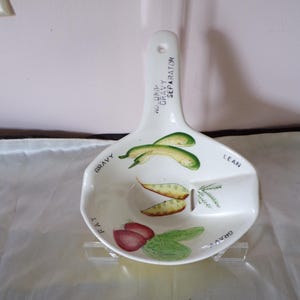 Op de afbeelding: Witte keramische jusafscheider met een handvat. De kom is verdeeld in secties met de labels "FAT", "LEAN" en "GRAVY", met geschilderde illustraties van groenten. Op het handvat staat "NO DRIP GRAVY SEPARATOR".